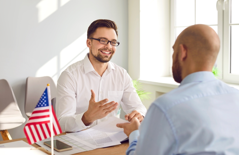 Happy, smiling, friendly young male Consul of United States of America sitting at office table with American flag