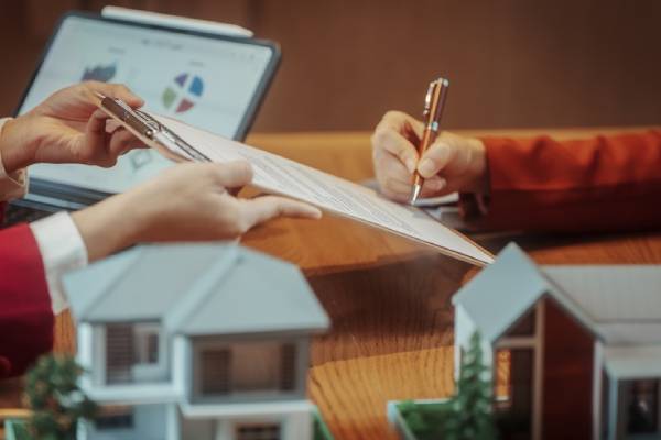 Hands signing and reviewing real estate contract at desk with laptop charts and house models